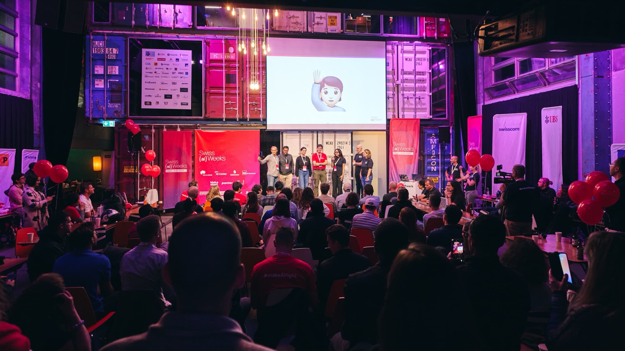 Crowd watching a team present on stage under red balloons